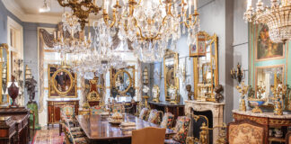 Inside Keil’s Antiques, display rooms show off breathtaking collectibles, including this long dining table framed by embroidered chairs and set below grand chandeliers.