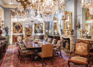 Inside Keil’s Antiques, display rooms show off breathtaking collectibles, including this long dining table framed by embroidered chairs and set below grand chandeliers.