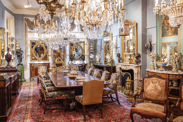 Inside Keil’s Antiques, display rooms show off breathtaking collectibles, including this long dining table framed by embroidered chairs and set below grand chandeliers.