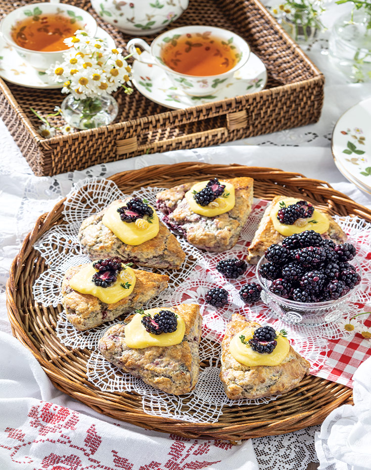 Blackberry-Thyme Scones nestle around a bowl of fresh blackberries. The pastries rest on a whicker tray, dappled in lace and red gingham.