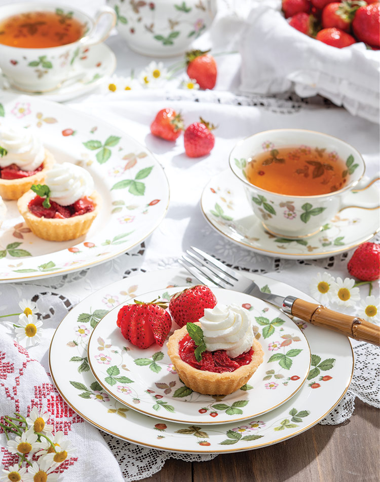 Miniature strawberry pies, sporting a coif of whipped cream and mint, huddle on a china plate beside a basket of fresh strawberries and two cups of tea.