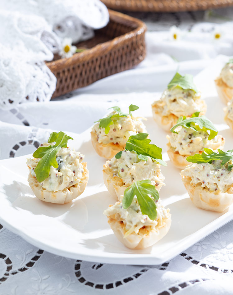 Mustard Chicken Salad Cups, accessorized in green arugula leaves, huddle on white plate, resting on a sunny painted white tablecloth.