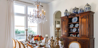 Kristie Shelton’s dining room features a large wooden table set for a harvest meal and adorned with pumpkin-colored roses. Floor-to-ceiling windows and a bespoke chandelier brighten this often-used room.
