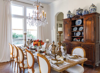 Kristie Shelton’s dining room features a large wooden table set for a harvest meal and adorned with pumpkin-colored roses. Floor-to-ceiling windows and a bespoke chandelier brighten this often-used room.