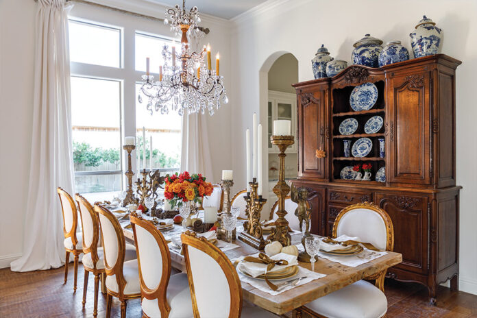 Kristie Shelton’s dining room features a large wooden table set for a harvest meal and adorned with pumpkin-colored roses. Floor-to-ceiling windows and a bespoke chandelier brighten this often-used room.