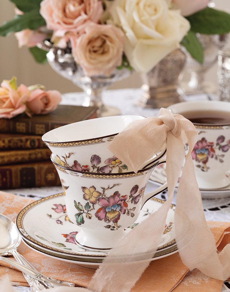 Blush roses grace a table set with feminine flair, where a stack of teacups tied with a ribbon await guests’ arrival.