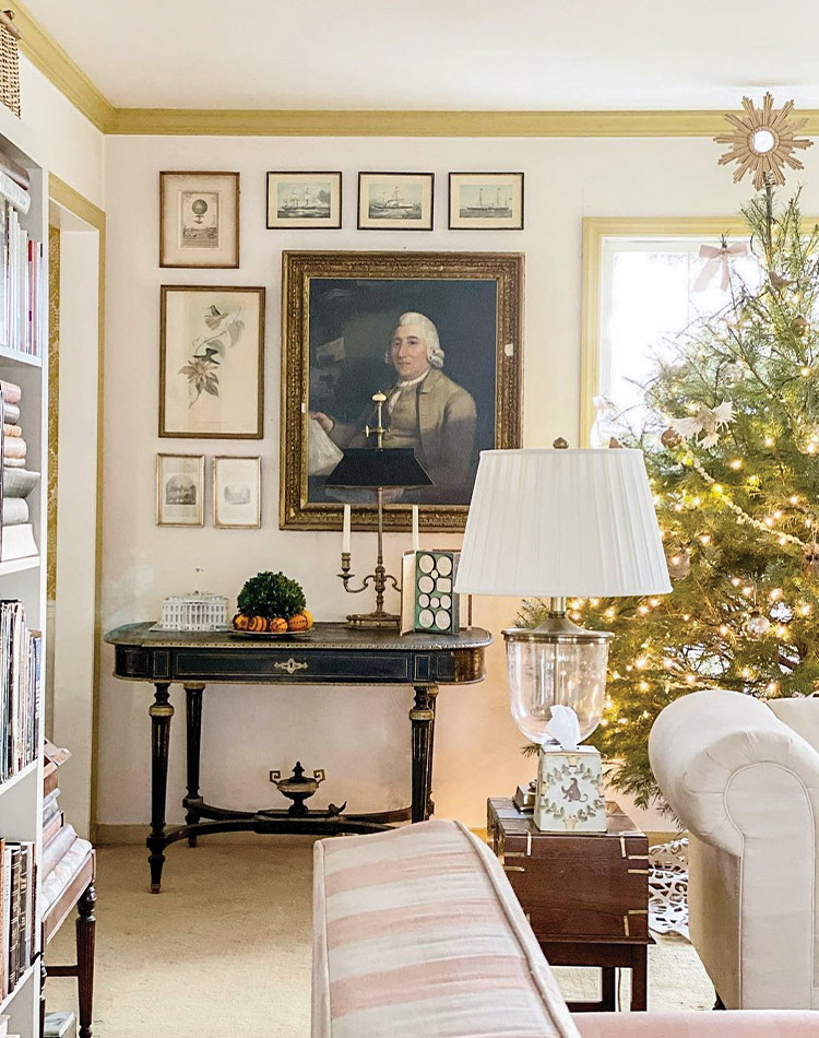 A corner of Jenny Bohannon’s living room shows a Christmas Tree with a sunburst tree topper and twinkly lights next to a black side table with a gallery wall of artwork hanging on the wall above.