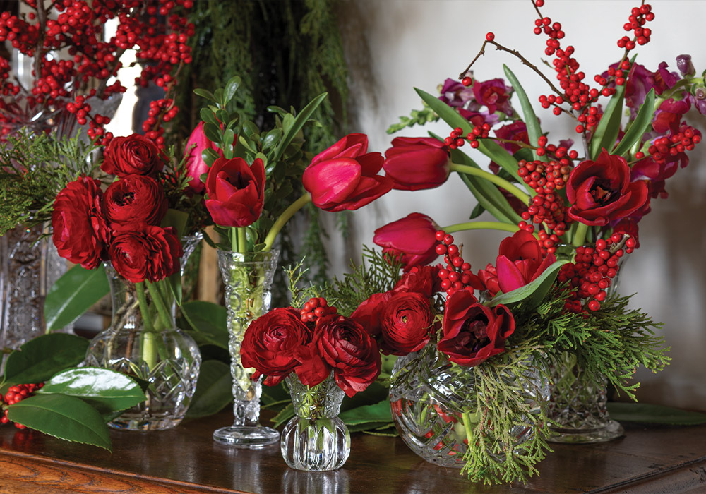 A sideboard table holds a collection of crystal vases in various shapes and sizes; each one cradles festive red berries and blooms.