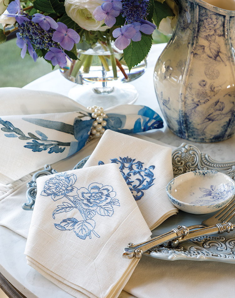 Blue Arte Italica plate with mixed blue embroidered Crown Linen Designs Napkins, pearl napkin ring, and platinum silver flatware aside a Giulietta blue patterned vase filled with purple and white flowers.