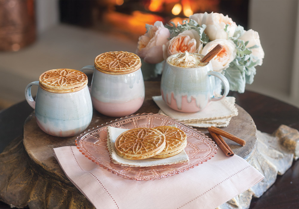 A trio of cozy blue and pink mugs filled with Spiced White Hot Chocolate are paired with sugary Stroopwafels atop a pink glass plate.