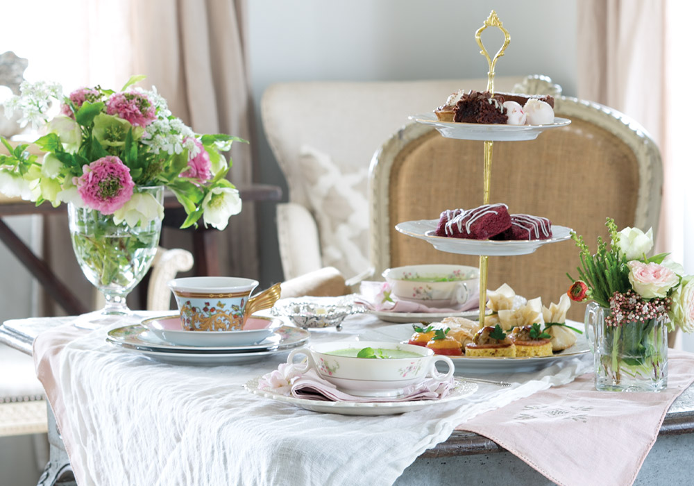 A table is set for tea with delicate porcelain cups, cheerful bouquets of pink and white flowers, and a tiered stand laden with red velvet scones, chocolate morsels, and dainty treats.