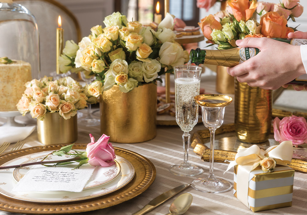 A lavish table is set with gold chargers topped with ivory China plates rimmed with gold and a crystal glass sparkles as Champagne is poured. An abundance of soft pink, white, and peach colored roses brim from gold vessels while candles flicker and a petite cake awaits beneath the clear glass dome of a cloche.