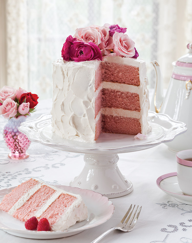 A pink velvet cake with vanilla Swiss meringue buttercream is topped by pink-hued roses and sits atop an ivory cake stand.