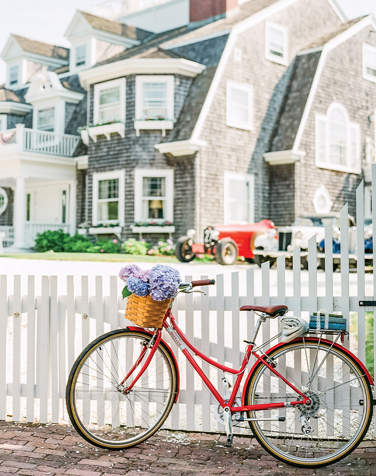 A red bicycle, with hydrangeas brimming from its forward basket and books secured to the rear, leans against the white picket fence of a classic Nantucket abode.