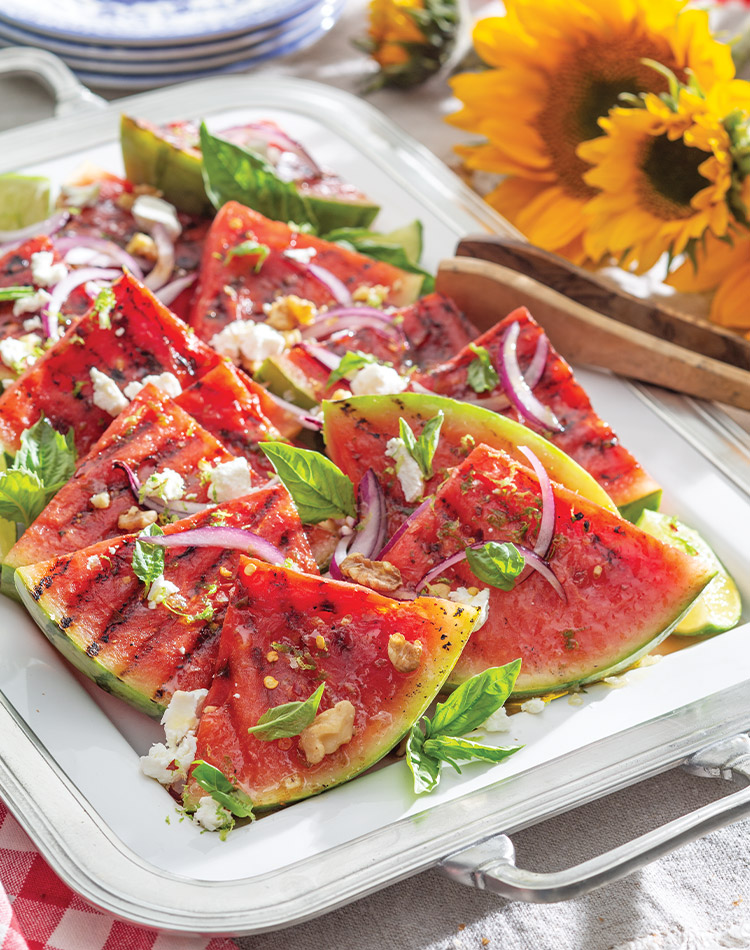 Grilled watermelon slices shingled on a serving platter with a silver handle, sits on a table beside sunflowers.