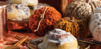 Atop a stack of autumnal china, a single cinnamon roll sits, while knit pumpkin décor looks on from the background.