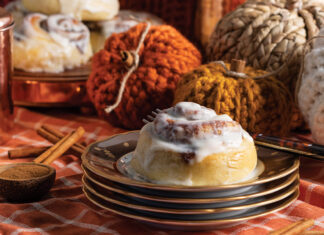 Atop a stack of autumnal china, a single cinnamon roll sits, while knit pumpkin décor looks on from the background.