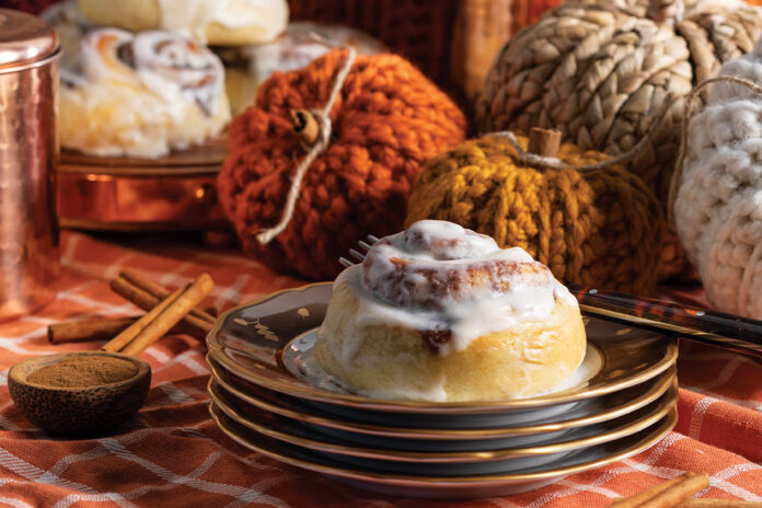Atop a stack of autumnal china, a single cinnamon roll sits, while knit pumpkin décor looks on from the background.