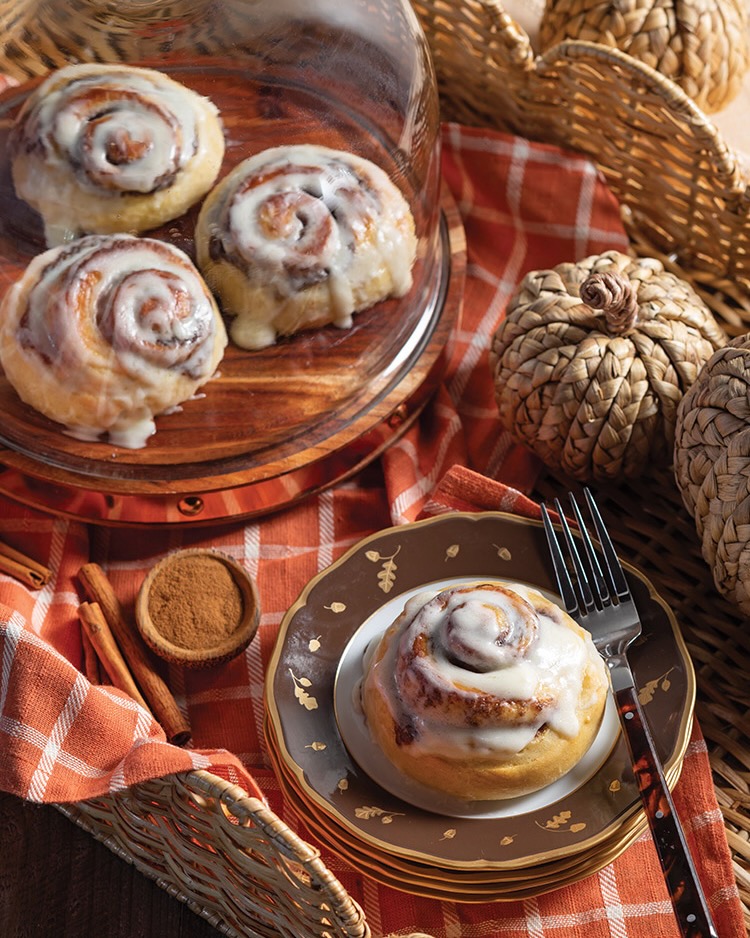 A trio of cinnamon rolls hides beneath a glass cake stand, while a single serving waits nearby on an autumnal stack of plates.