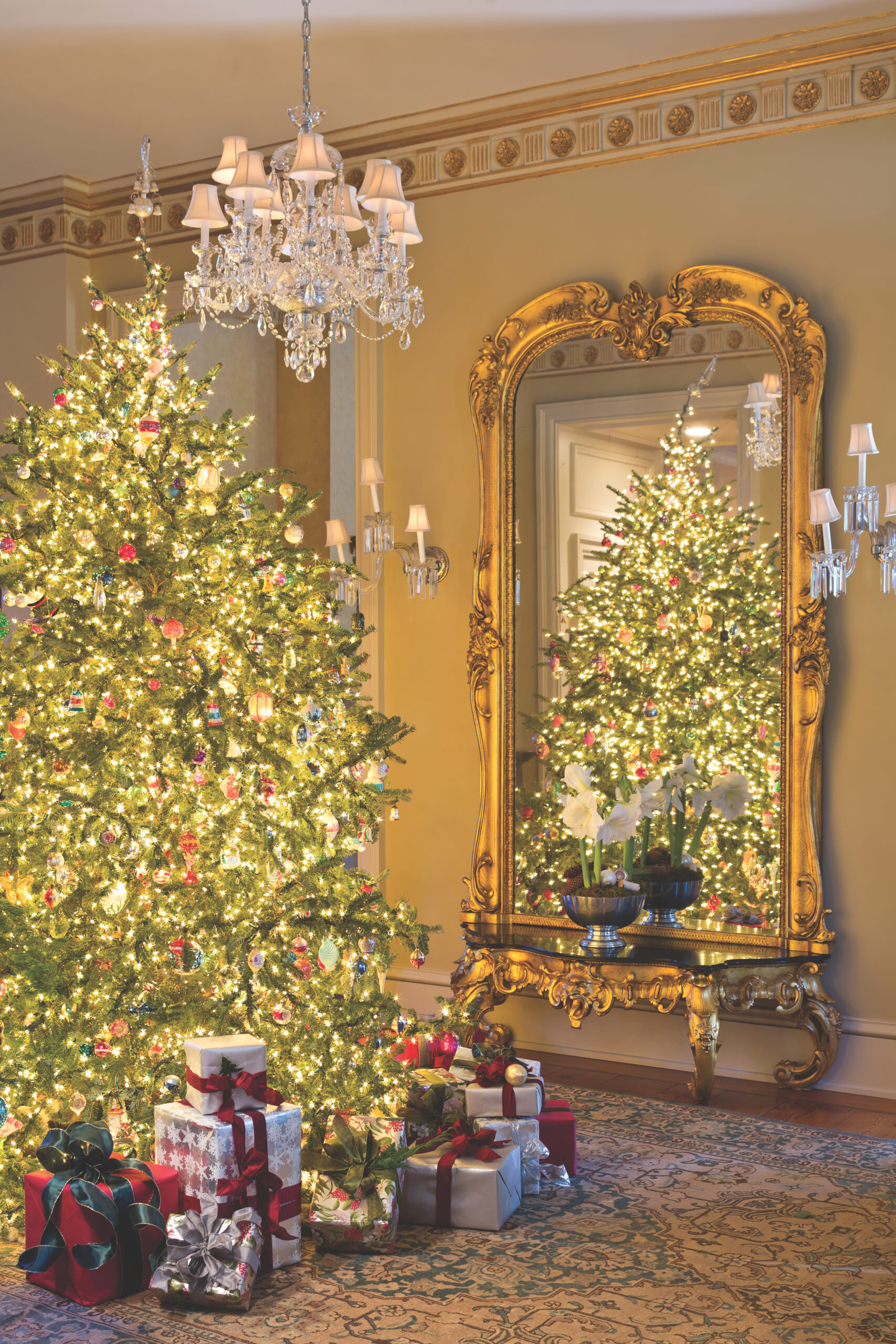 A room adorned with a gold wallpaper border, a chandelier, an ornate gold mirror, and a Christmas tree surrounded by gifts.