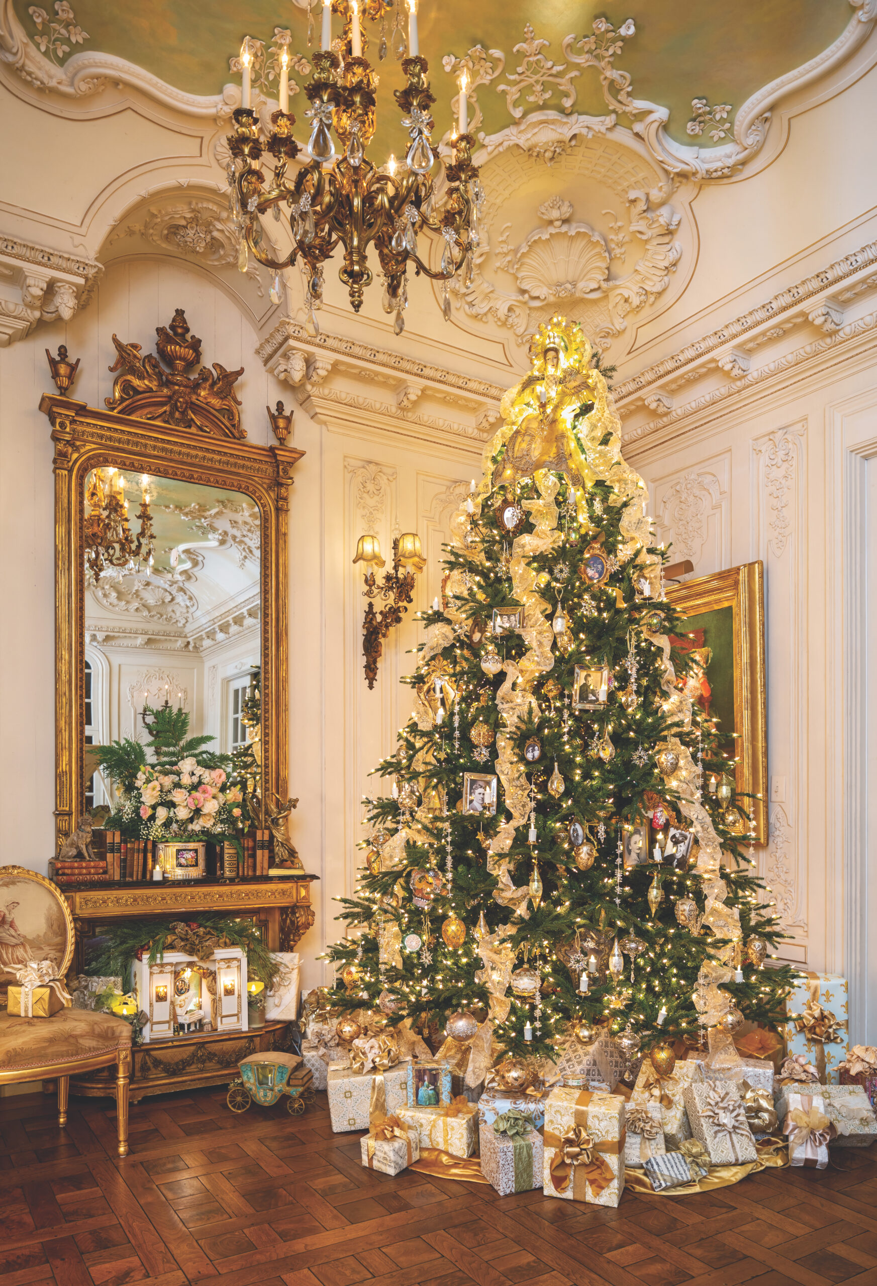 A Christmas tree decorated in a cream palette, sitting in a lavish room adorned with ornate millwork, a chandelier, and a gold mirror.