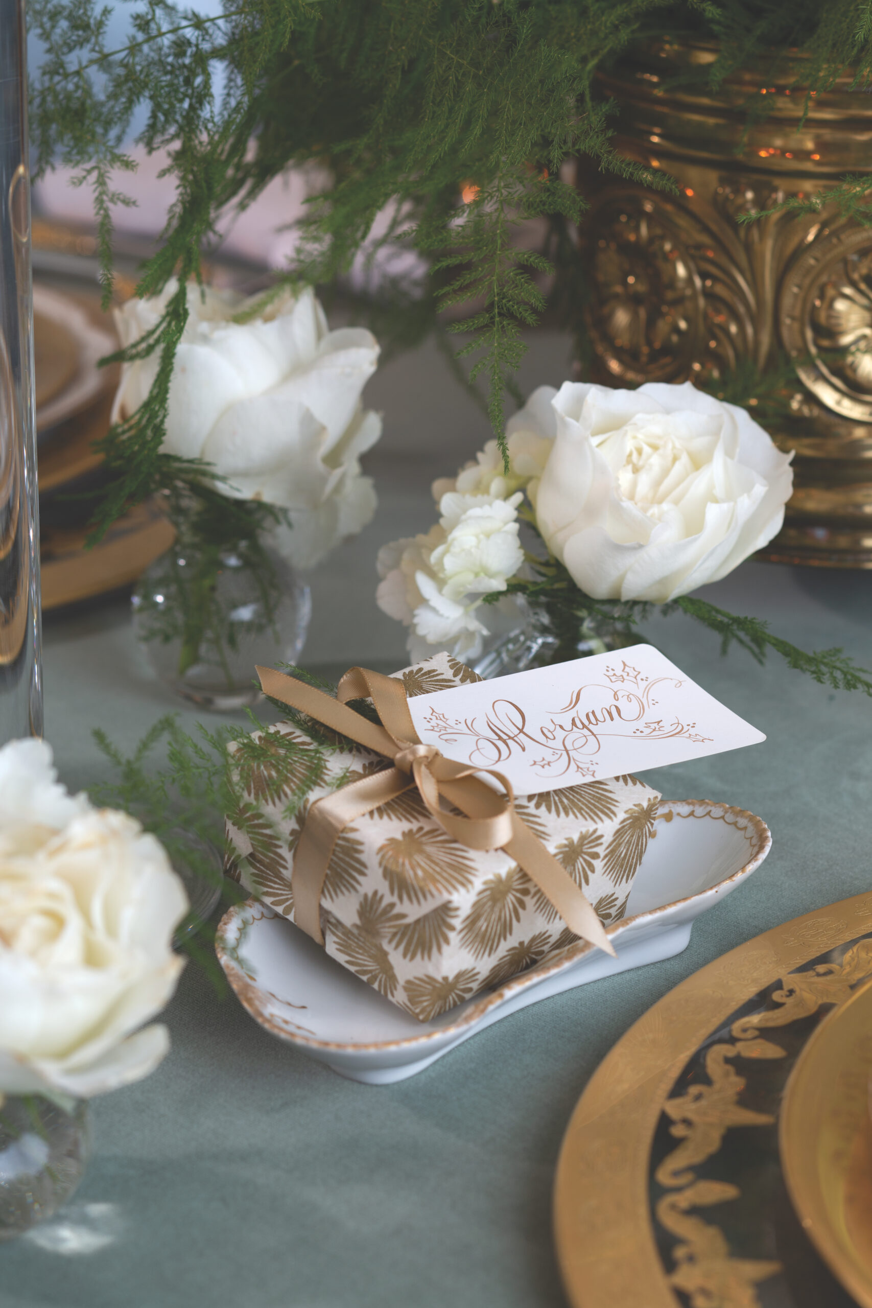A tiny gift wrapped in gold-and-white patterned paper and tagged with a calligraphed name tag, surrounded by white roses on a table.