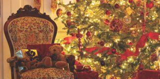 A Christmas tree decorated with red ornaments and ribbons is aglow and surrounded by a wreath, an antique chair, and wrapped gifts.