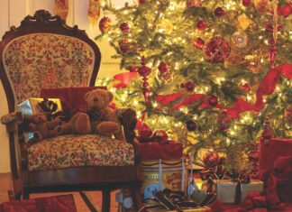A Christmas tree decorated with red ornaments and ribbons is aglow and surrounded by a wreath, an antique chair, and wrapped gifts.