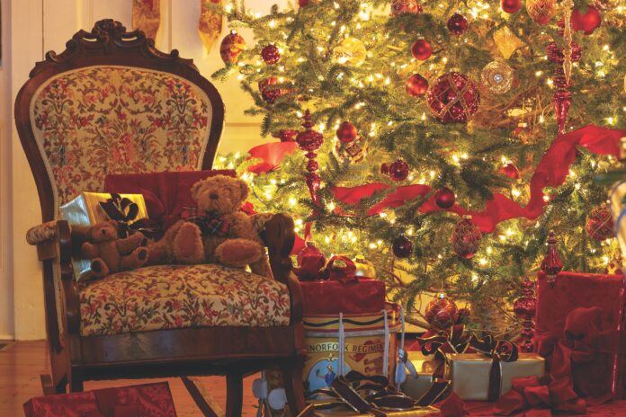 A Christmas tree decorated with red ornaments and ribbons is aglow and surrounded by a wreath, an antique chair, and wrapped gifts.