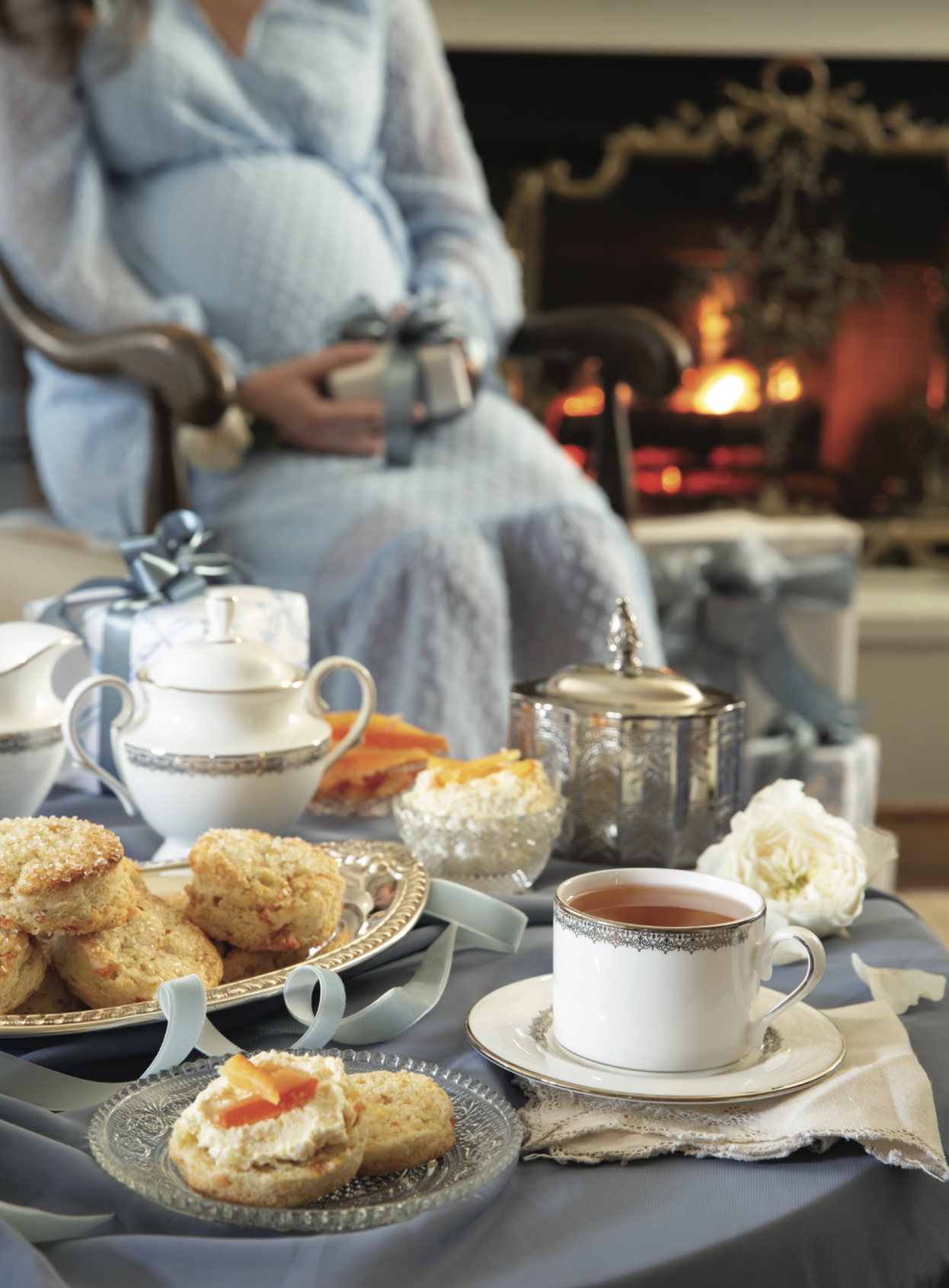 A pregnant woman sits in the background near a fireplace, with a tea spread arranged in the foreground.