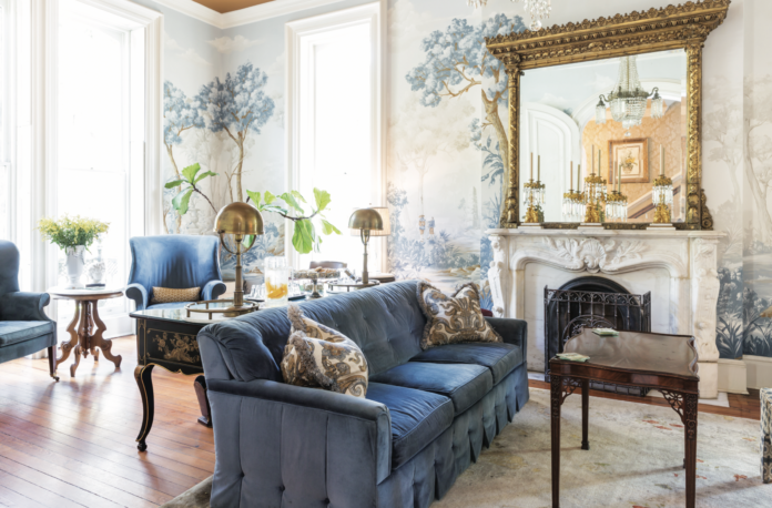 The luxurious antique interior of Savannah's Hamilton-Turner Inn, featuring a sofa with blue upholstery, a wallpaper mural, a fireplace, and a gilfed mirror.