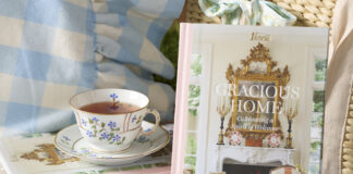 A stack of books with a pink spine are crowned with a porcelain cup of tea, beside which the cover of Gracious Home is displayed.