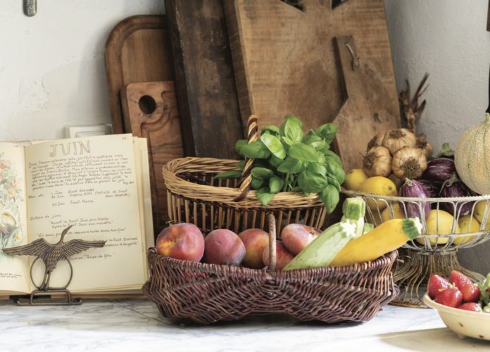 A counter in a French Country kitchen with cutting boards, a cookbook, and baskets of fresh vegetables.