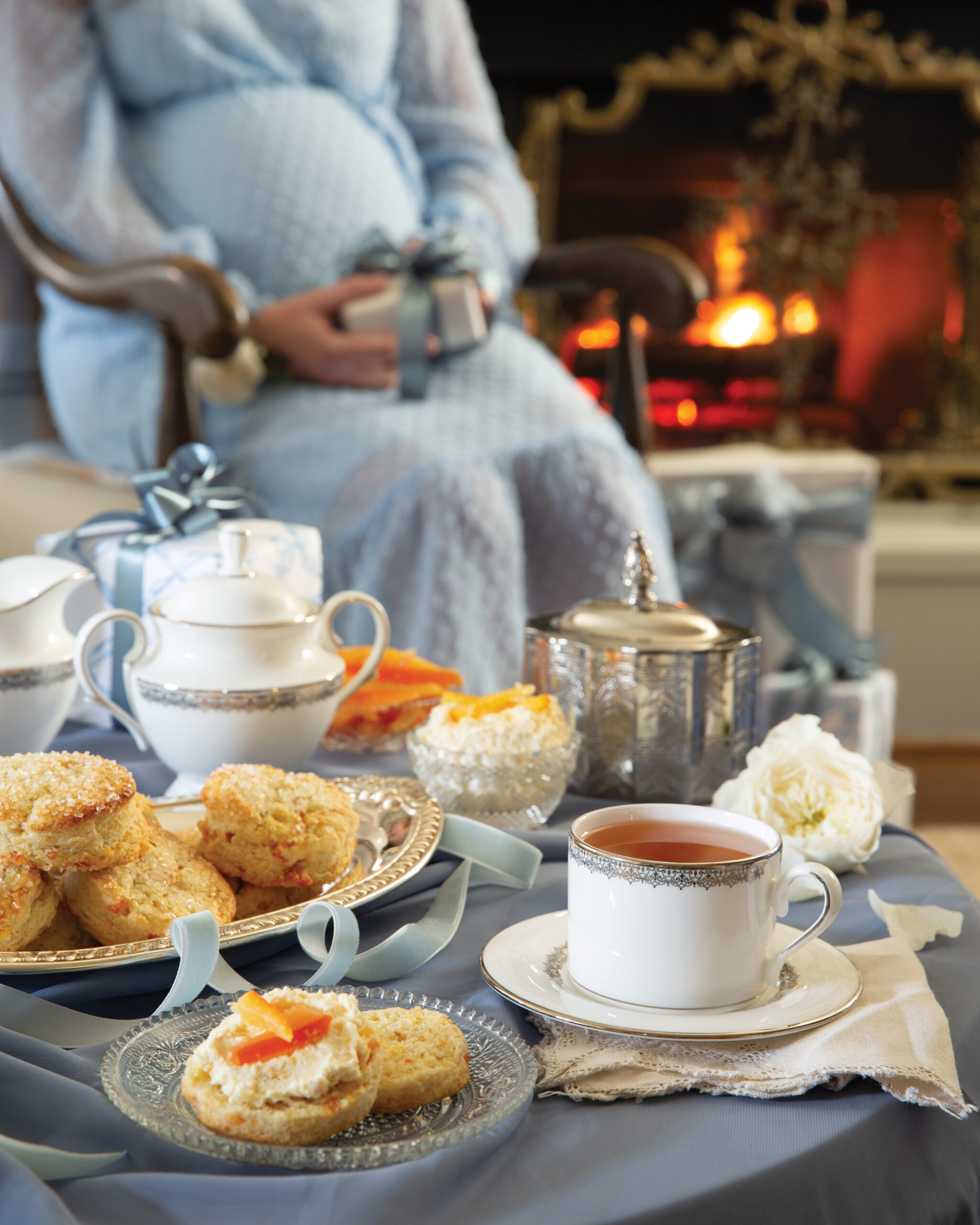 Table with scones, clotted cream, and tea in front of pregnant woman sitting in front of fireplace 