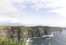 The Cliffs of Moher rise dramatically above the Atlantic Ocean, their sheer green-topped rock faces plunging into crashing waves below beneath a wide Irish sky.