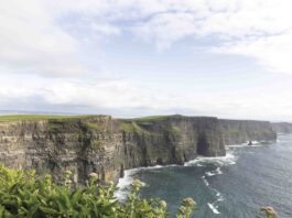 The Cliffs of Moher rise dramatically above the Atlantic Ocean, their sheer green-topped rock faces plunging into crashing waves below beneath a wide Irish sky.