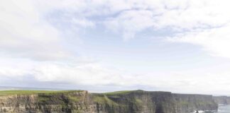 The Cliffs of Moher rise dramatically above the Atlantic Ocean, their sheer green-topped rock faces plunging into crashing waves below beneath a wide Irish sky.