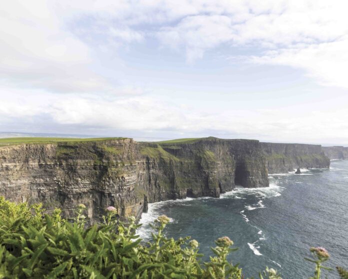 The Cliffs of Moher rise dramatically above the Atlantic Ocean, their sheer green-topped rock faces plunging into crashing waves below beneath a wide Irish sky.