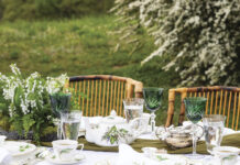Elegant spring tablescape set outdoors with lily-of-the-valley china, green crystal goblets, silver tea service, and a floral centerpiece on a white tablecloth surrounded by wicker chairs and blooming garden greenery.