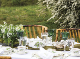 Elegant spring tablescape set outdoors with lily-of-the-valley china, green crystal goblets, silver tea service, and a floral centerpiece on a white tablecloth surrounded by wicker chairs and blooming garden greenery.