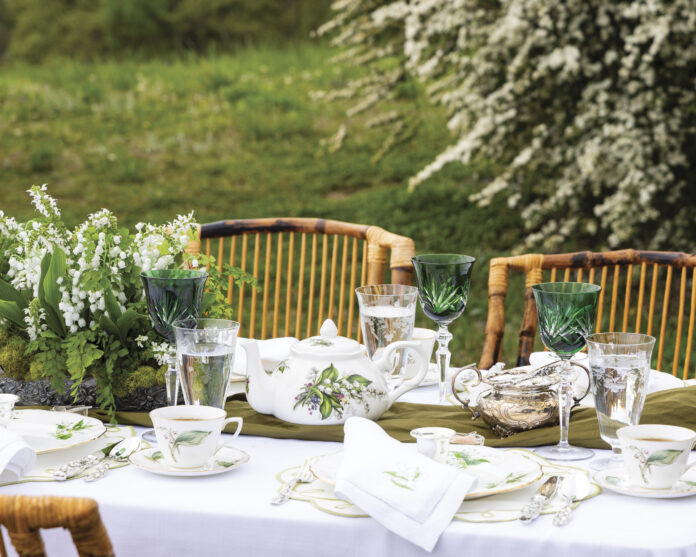 Elegant spring tablescape set outdoors with lily-of-the-valley china, green crystal goblets, silver tea service, and a floral centerpiece on a white tablecloth surrounded by wicker chairs and blooming garden greenery.