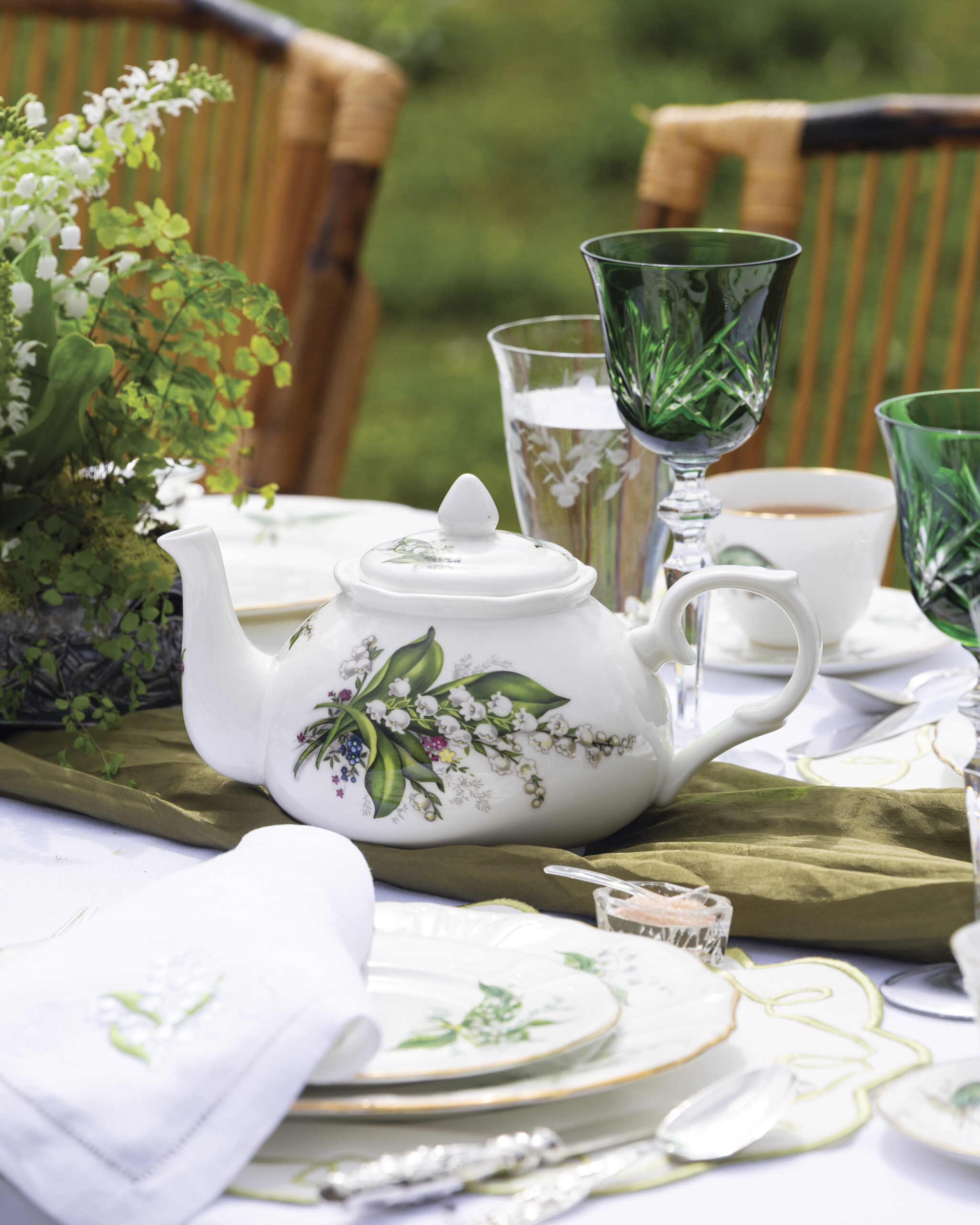 Alt text: Lily-of-the-valley teapot displayed on an elegant outdoor spring table setting with green crystal goblets, floral china, silver flatware, and garden greenery in the background.