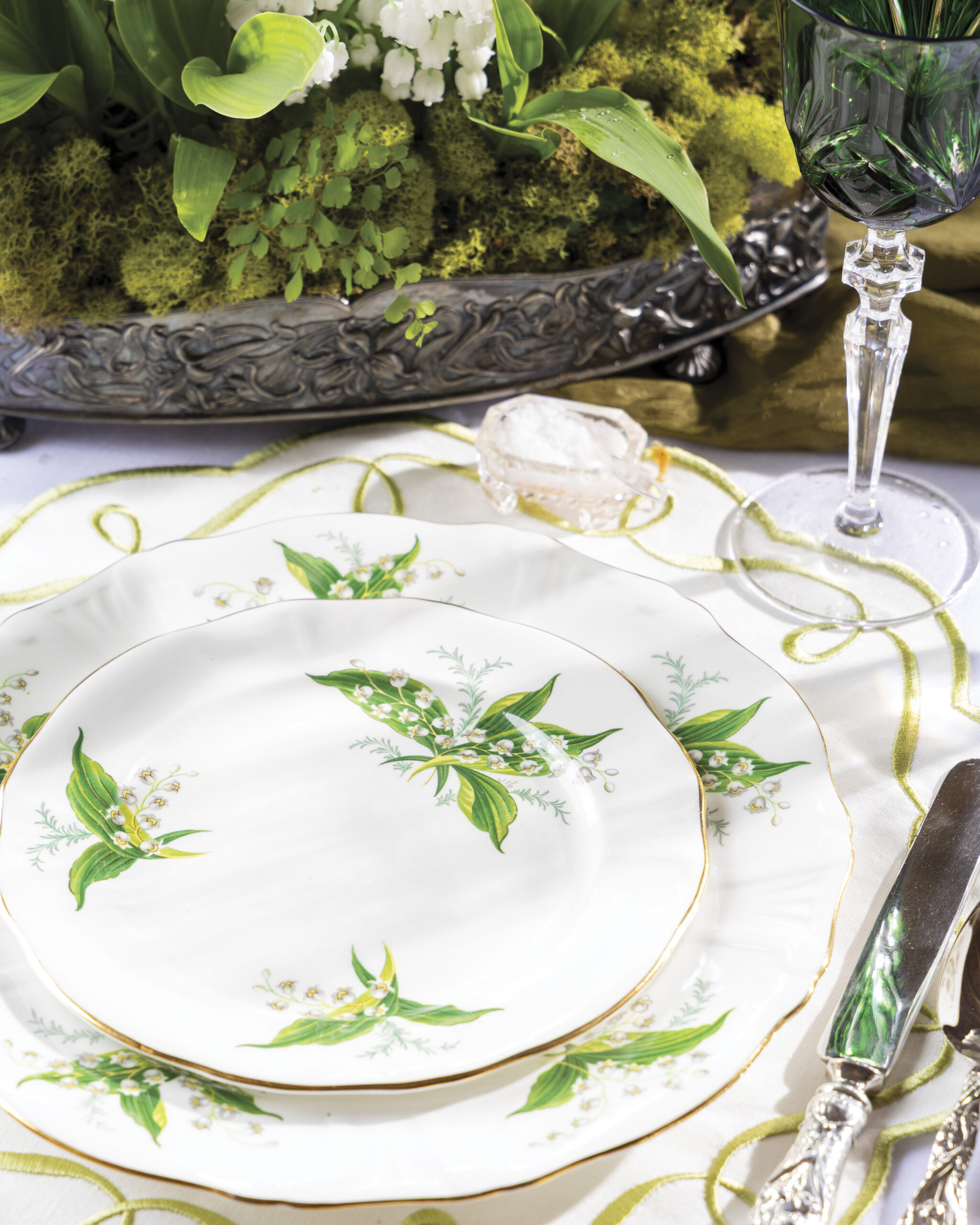 Close-up of lily-of-the-valley patterned china plates with gold trim arranged on a white tablecloth, accompanied by silver flatware, a green crystal goblet, and a moss and floral centerpiece.