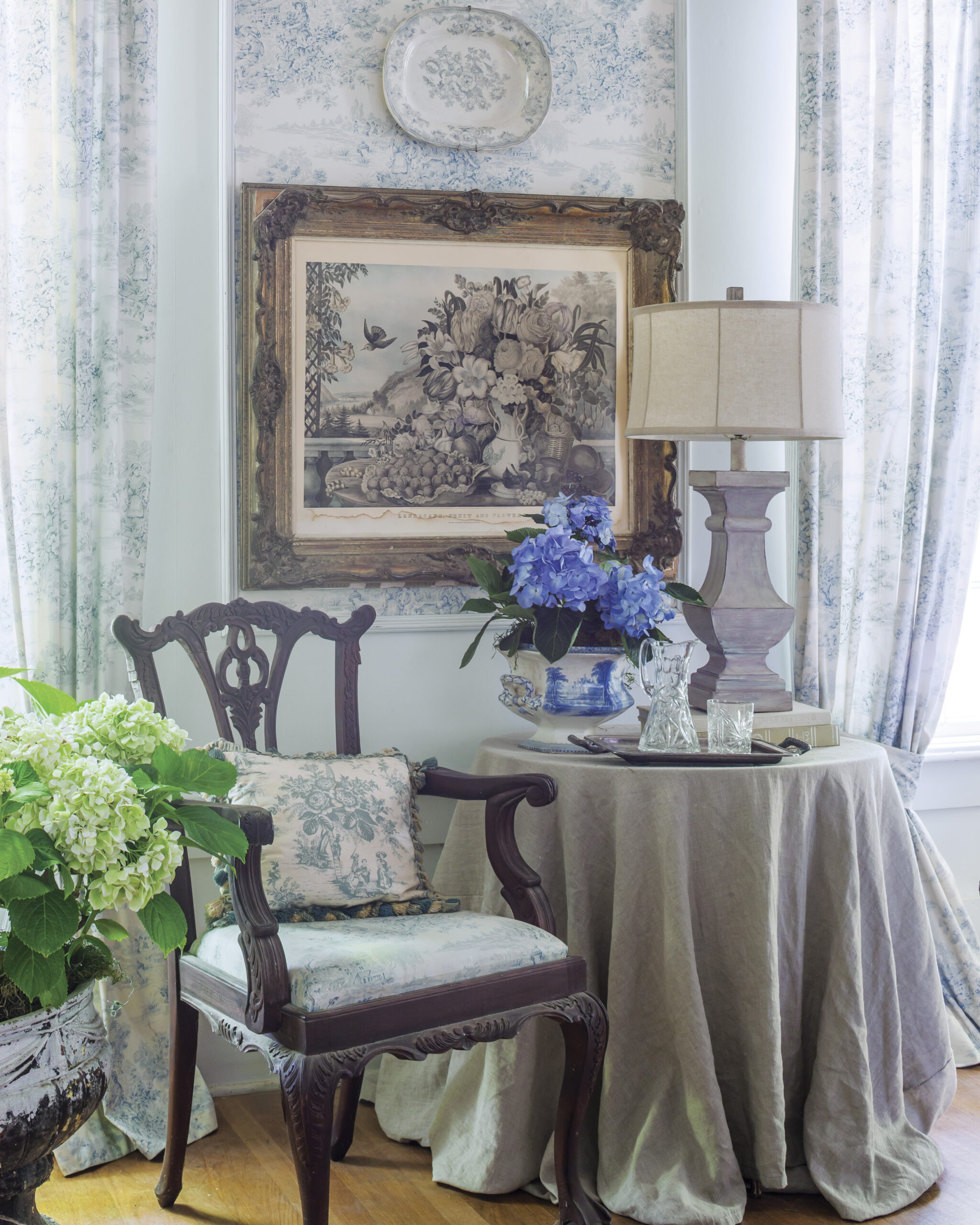 A softly lit sitting corner with blue-and-white toile wallpaper, an antique wooden chair with a patterned cushion, and a round table draped in linen holding a lamp, crystal glasses, and a vase of blue hydrangeas beneath a framed floral artwork.