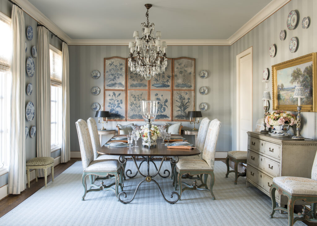 An elegant dining room with soft blue striped walls, a crystal chandelier, and a dark wood table surrounded by upholstered chairs, set with glassware and flowers. Blue-and-white plates and botanical panels decorate the walls, while a painted sideboard holds floral arrangements and lamps.