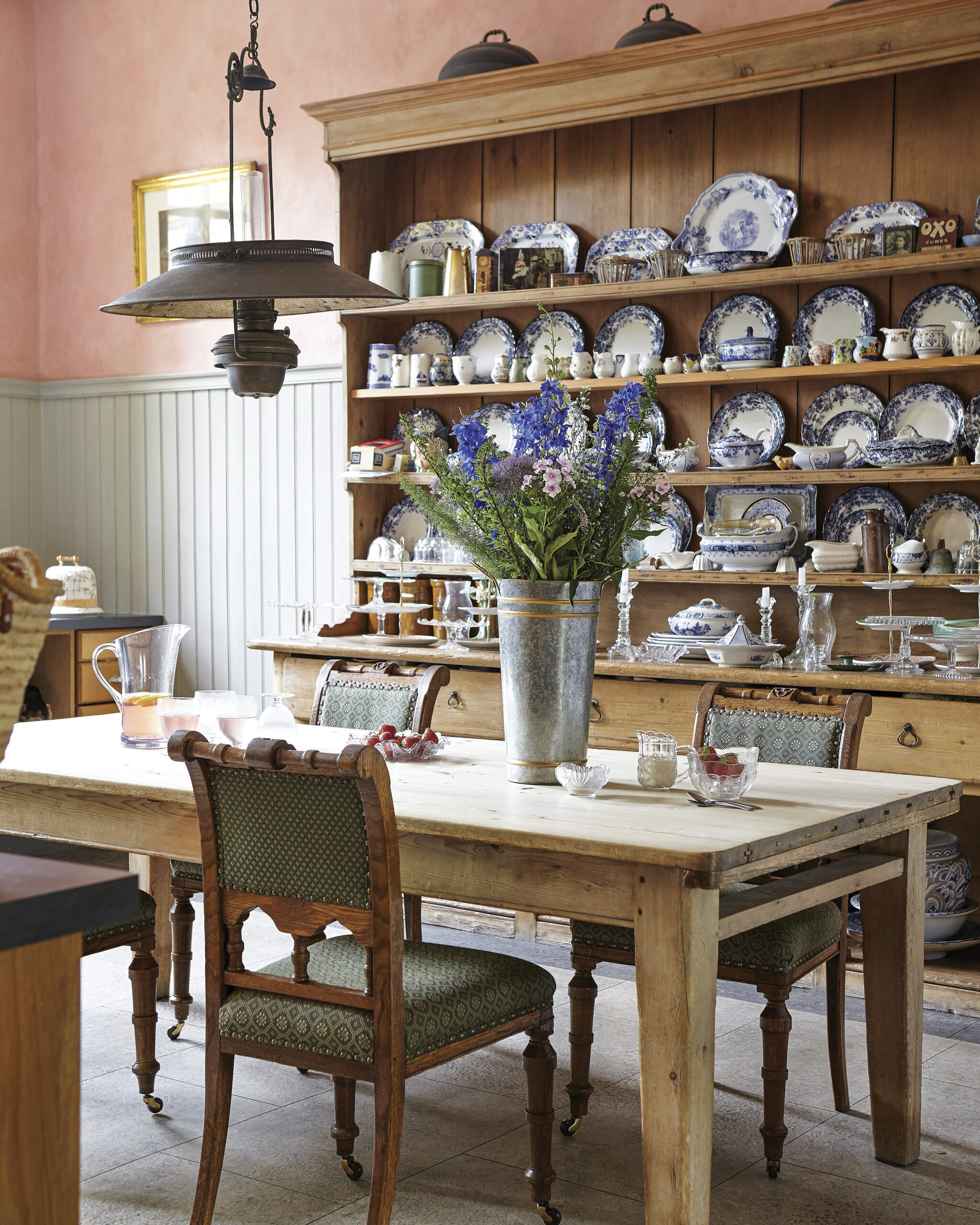 A rustic dining room with a wooden table and upholstered chairs, set with glassware and a bouquet of blue wildflowers. Behind it, a large wooden hutch displays blue-and-white china, while a metal pendant light hangs overhead against soft pink walls.