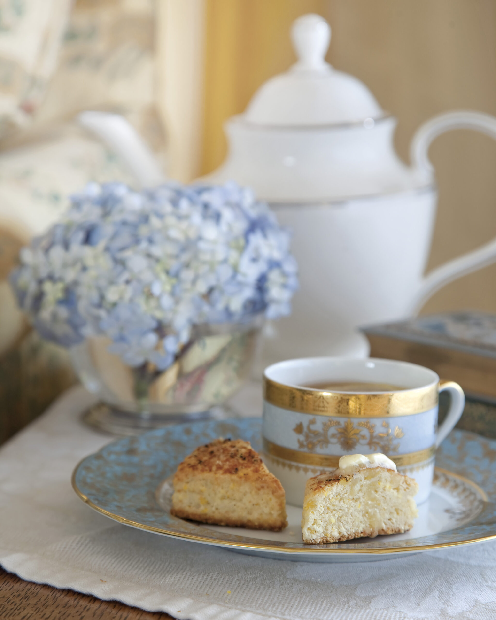 A delicate tea setting with a blue-and-gold china cup and saucer, served with a split scone topped with cream, set before a white teapot and a small arrangement of blue hydrangeas in soft focus.