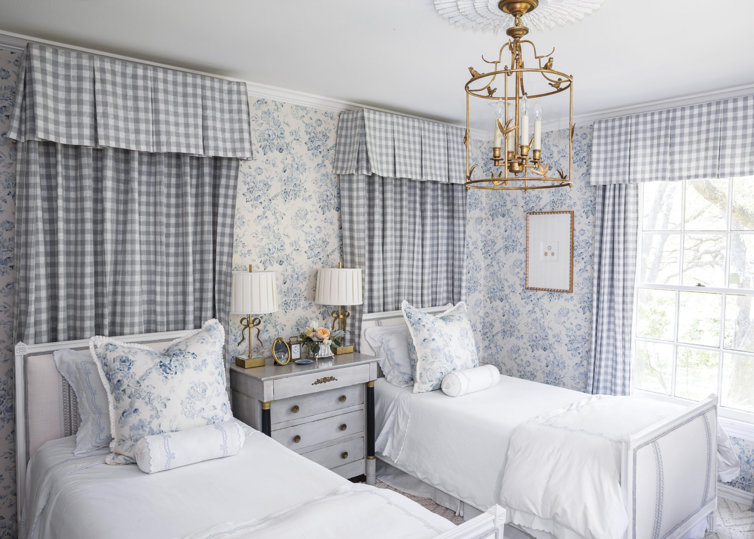A light-filled bedroom with two twin beds dressed in white linens and blue floral pillows, set against blue-and-white patterned wallpaper and gingham curtains. A painted nightstand with matching lamps sits between the beds, beneath a gold chandelier.
