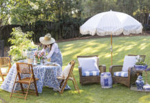 Woman in a straw hat arranging flowers on a blue-and-white table set for outdoor spring dining, with wicker chairs, striped umbrella, and garden seating on a sunny lawn.