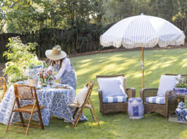 Woman in a straw hat arranging flowers on a blue-and-white table set for outdoor spring dining, with wicker chairs, striped umbrella, and garden seating on a sunny lawn.
